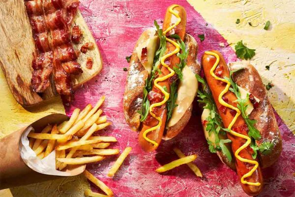 2 Hot Dogs in a bun with chips and bacon aside on a coloured wooden board
